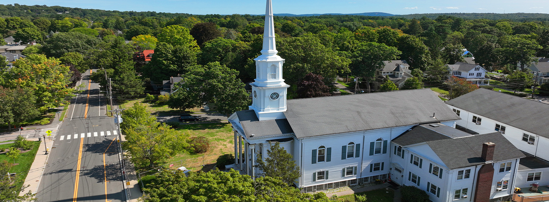 The image shows a church with a steeple prominently visible against a clear sky, surrounded by trees and grass, with a road leading up to it and a building with a red roof on its left side in the foreground.