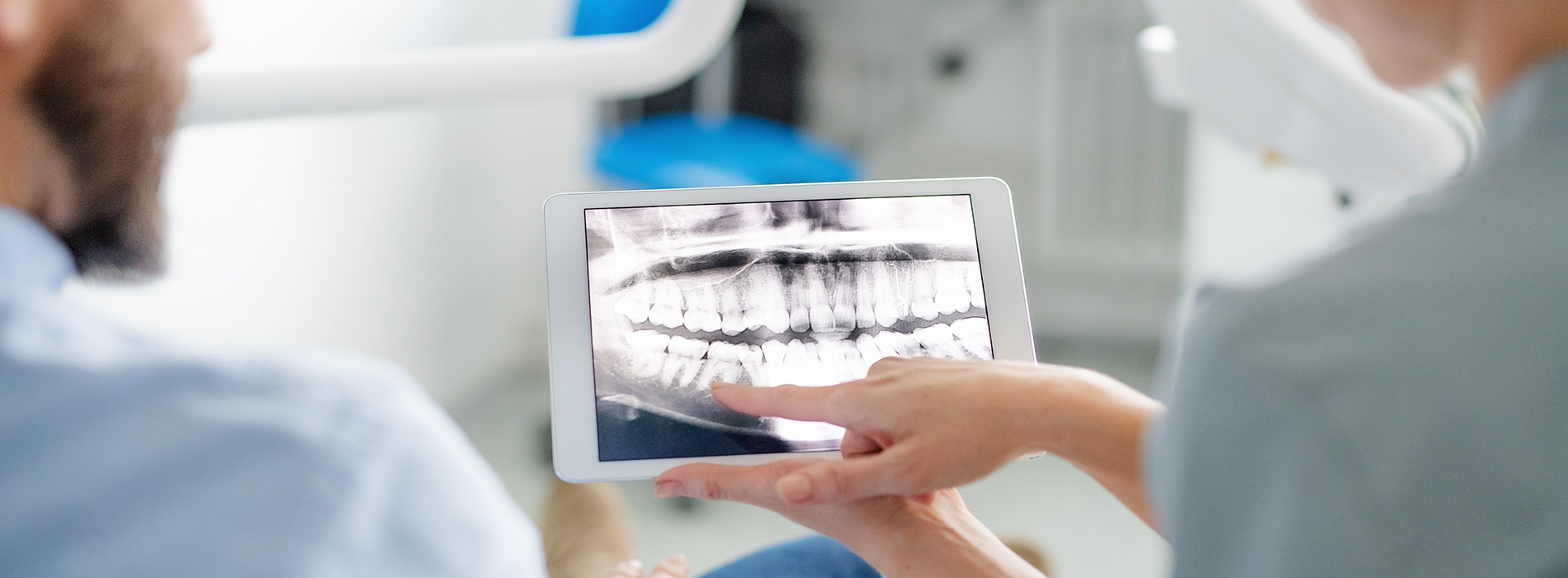 A dental professional holding up an X-ray on a tablet while seated at a workstation with another person present in a medical setting.