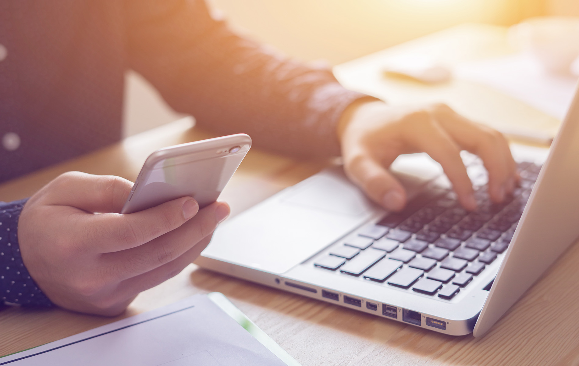 The image shows a person using a laptop with their hands while another person s hands are seen holding a smartphone, both set against a blurred background suggesting an office environment.