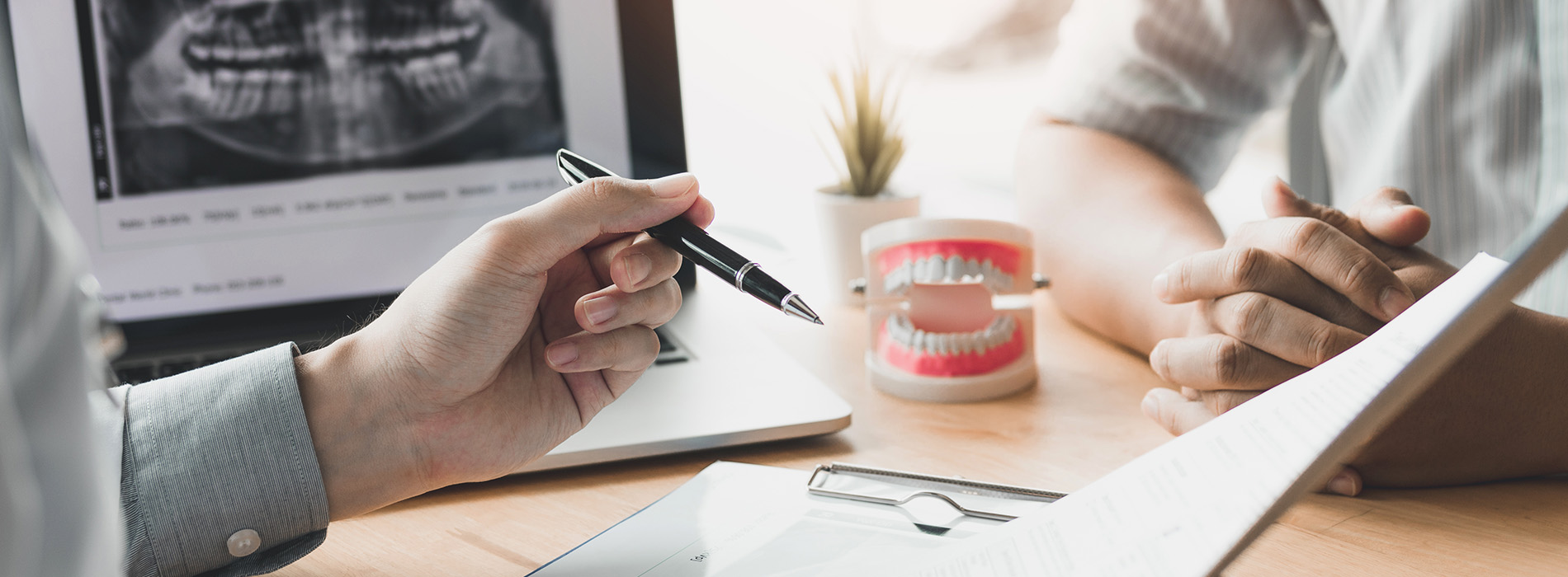 The image shows two hands holding pens over a table with a laptop, while in the background, there s a person sitting at another table with a computer monitor displaying medical images, and the scene suggests an office setting.